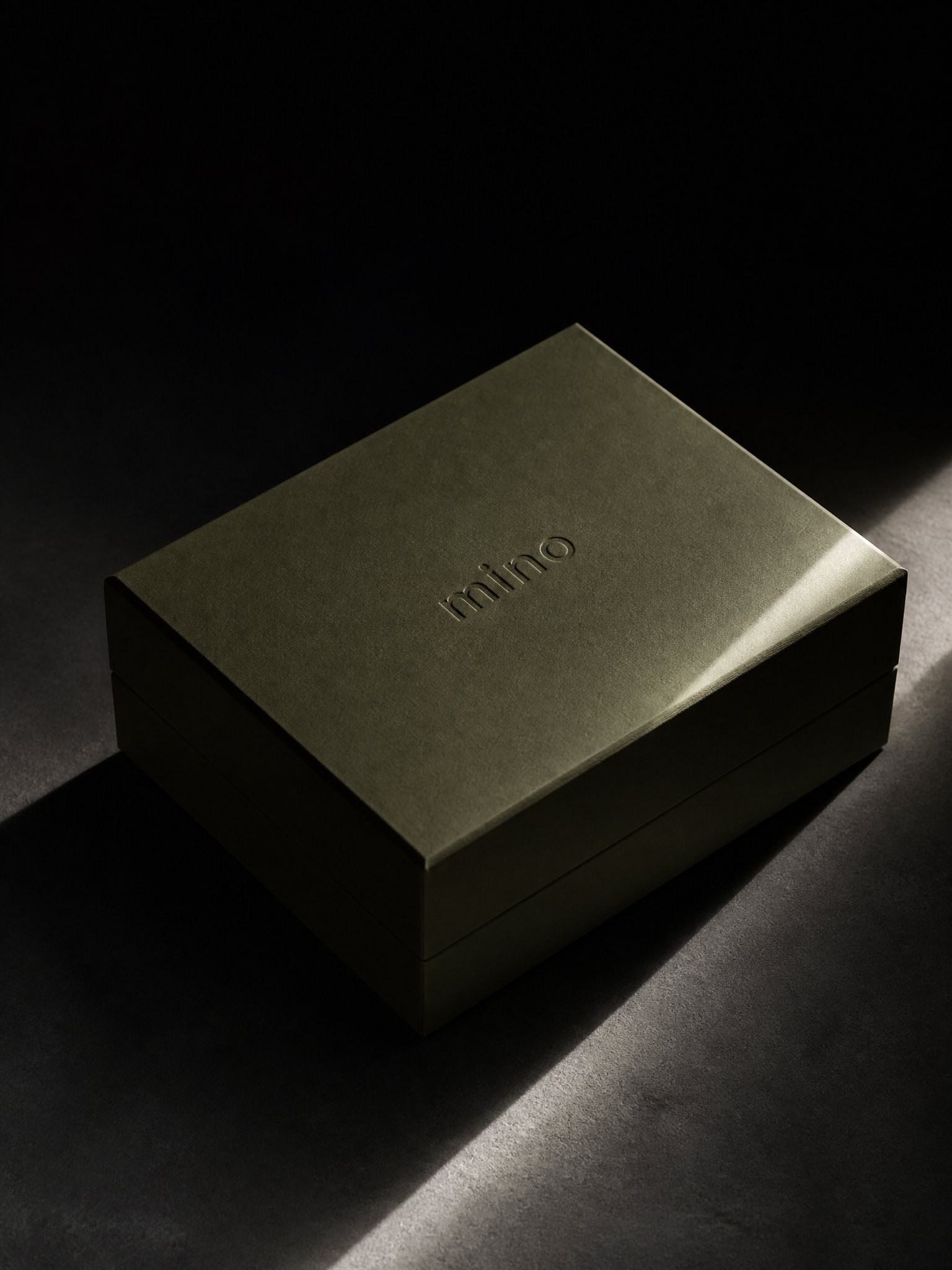 A closed sage gift box with a debossed mino logo, lit by a single hard beam